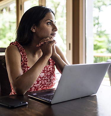 Woman at laptop with hands clasped stares out window