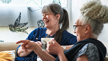 Two women laughing and drinking coffee in a living room setting.