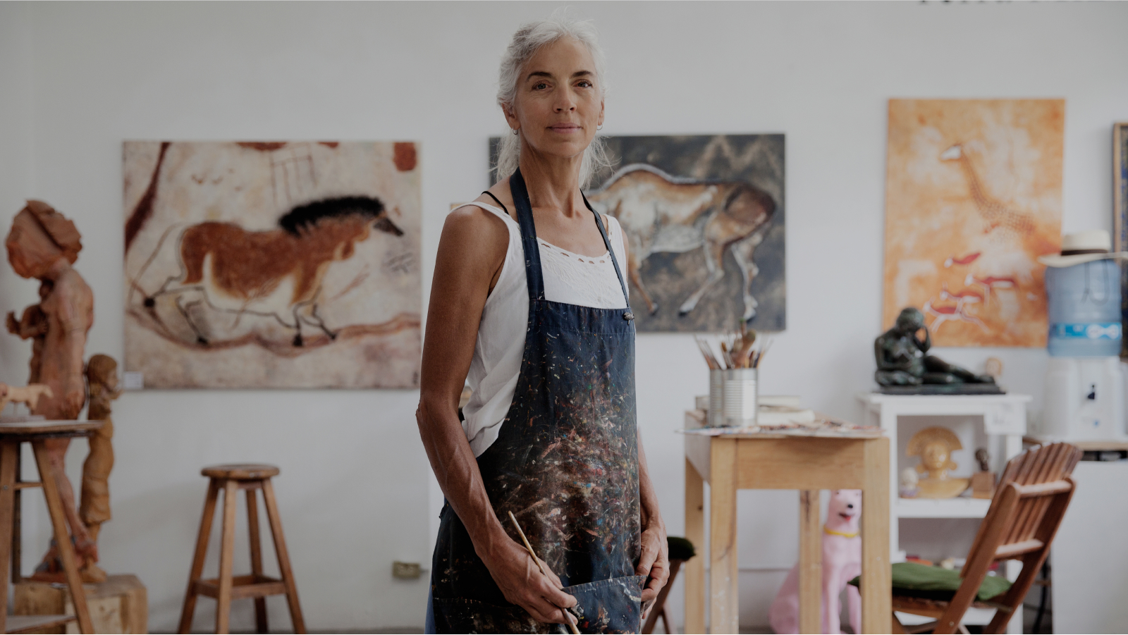 A woman in a paint-covered smock stands in a room, surrounded with paintings.