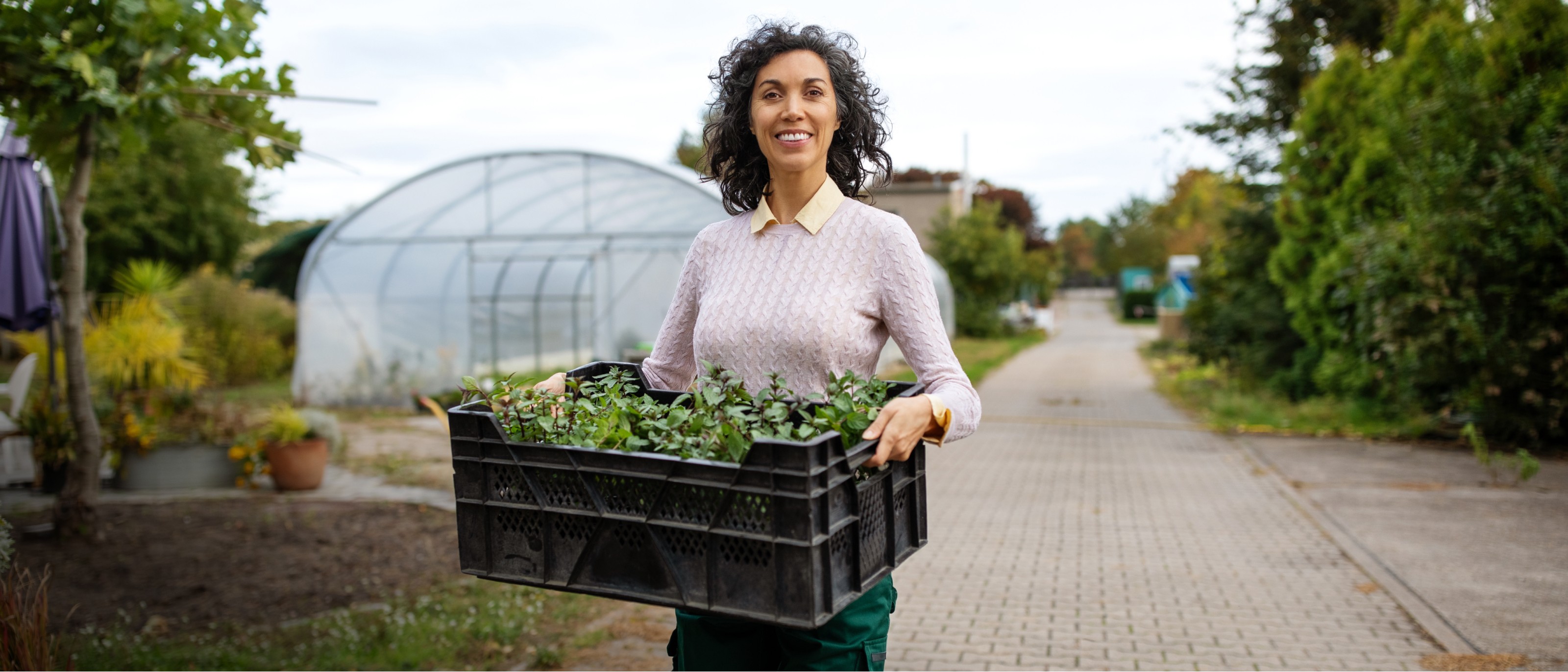 Woman smiling standing in front of greenhouse holding case of fresh plants